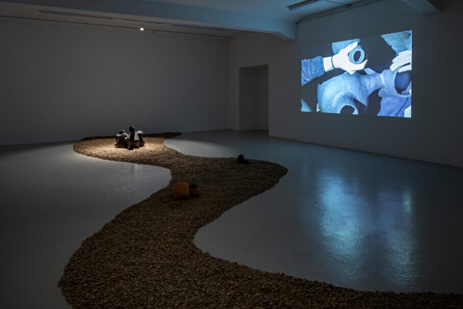 A winding path of gravel curves across a gallery floor, with figures seated along its route. A video projection on the right wall shows close-up footage of hands working with blue objects. The installation creates an immersive environment exploring ritualistic or meditative practices.
