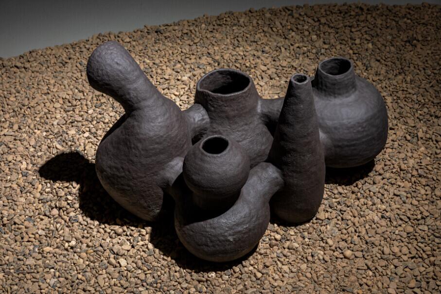 A collection of dark ceramic vessels with irregular, organic shapes arranged on a bed of tan gravel. The pots feature various curved and bulbous forms with openings of different sizes, creating an abstract, almost sculptural composition.