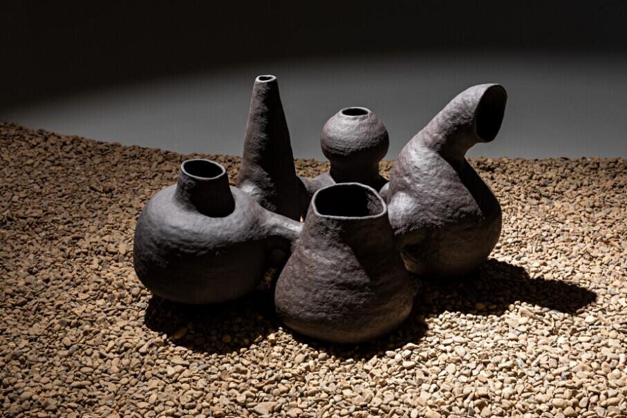 Five dark ceramic vessels with irregular, organic shapes and textured surfaces sit on sandy ground under dramatic lighting. The vessels feature various spout and opening designs, evoking ancient pottery forms. Their asymmetrical forms and weathered appearance suggest ritualistic or ceremonial purposes.