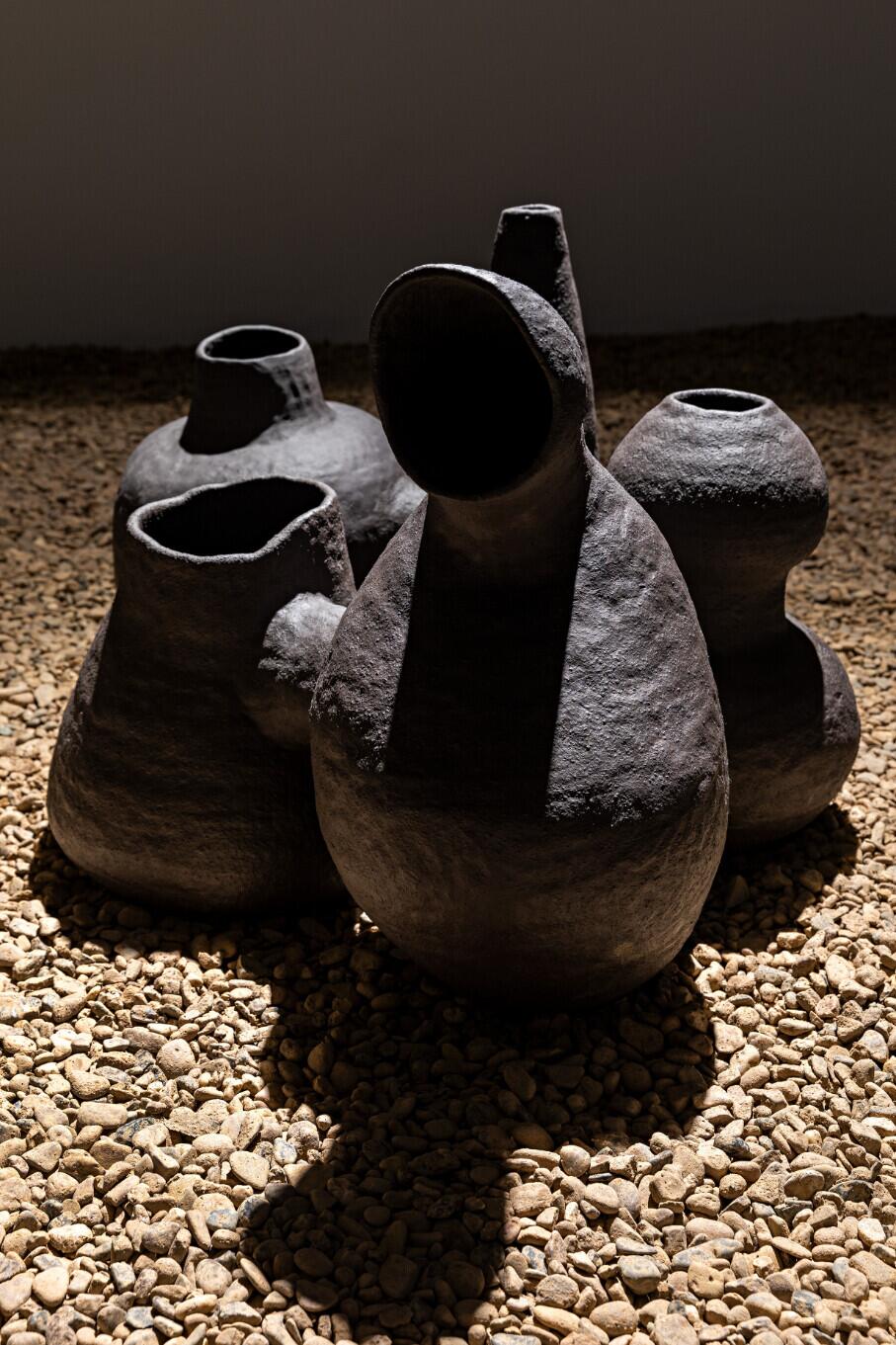 Five dark ceramic vessels of varying heights and shapes sit on a bed of small gravel against a dark background, their openings casting deep shadows. The vessels have rough, textured surfaces with lighter weathered edges around their rims. Strong directional lighting creates dramatic shadows that emphasize the sculptural forms of these primitive-looking clay pots.