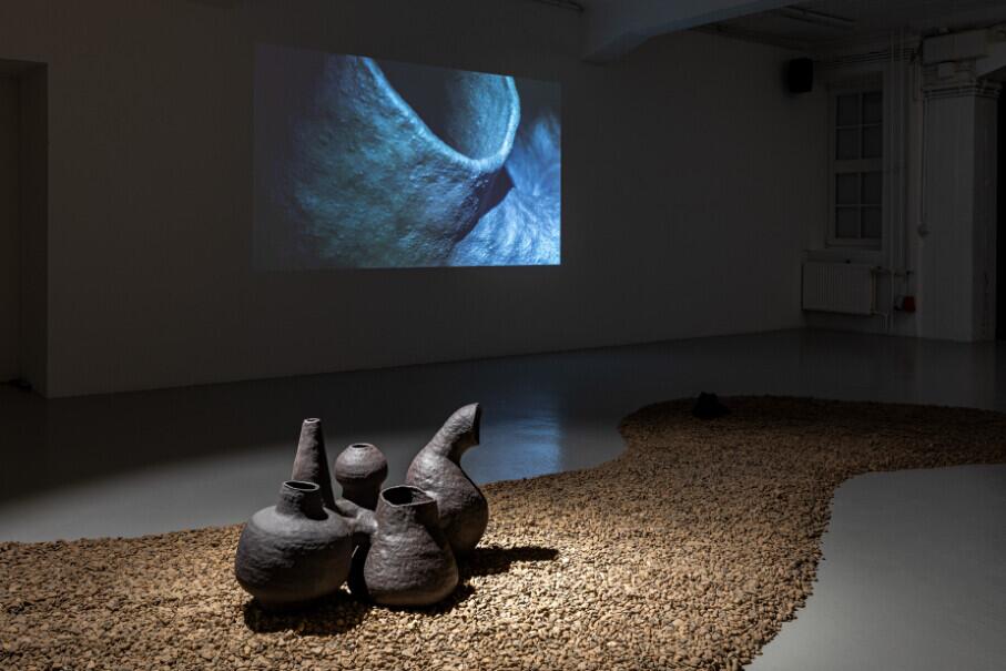 Two dark sculptural forms resembling crouching figures sit on a bed of gravel in a gallery space, facing a projection screen displaying an abstract blue and white moving image. The installation combines physical sculpture with projected video in a minimalist exhibition setting.
