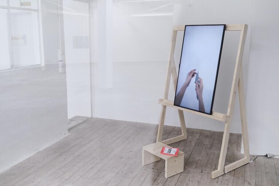 A wooden easel with a light wood frame stands in a minimalist white gallery space, displaying a vertical monitor showing a close-up of hands. Below the screen sits a small wooden shelf holding what appears to be printed materials in red and white.