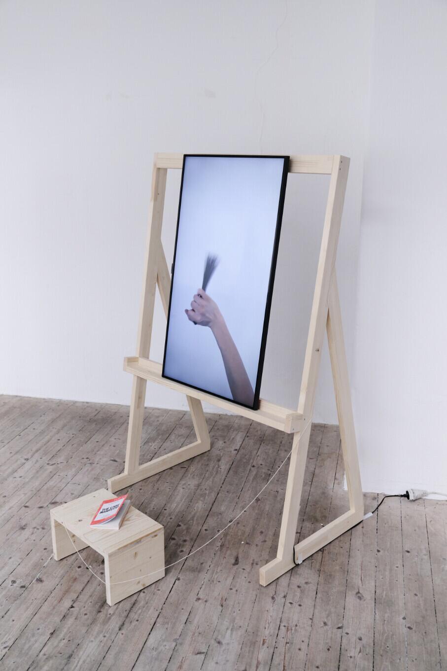 A wooden easel with a digital screen displays a hand holding a toothbrush, with a small wooden block positioned at its base on a wooden floor against a white wall.