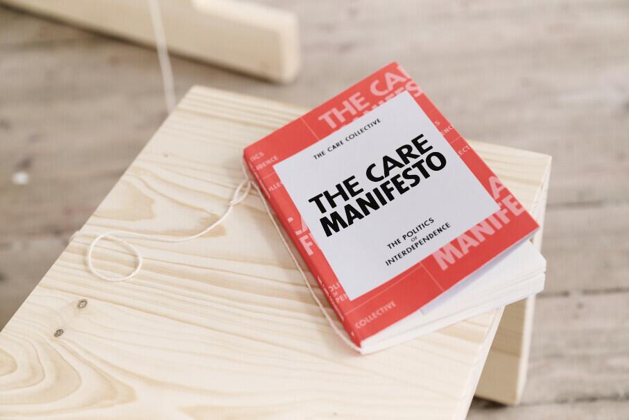 A red book titled “The Care Manifesto” lays on a light wooden bench.