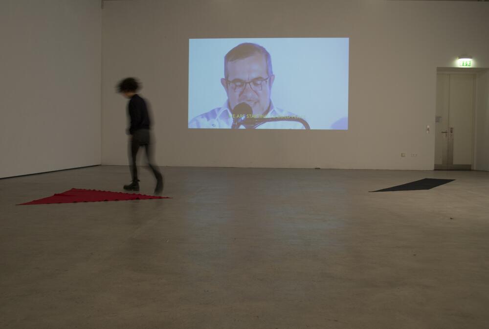 A person stands on a red carpet in a gallery space, watching a video projection on the white wall ahead. The projection displays a bald man wearing glasses and speaking into a microphone against a light blue background.