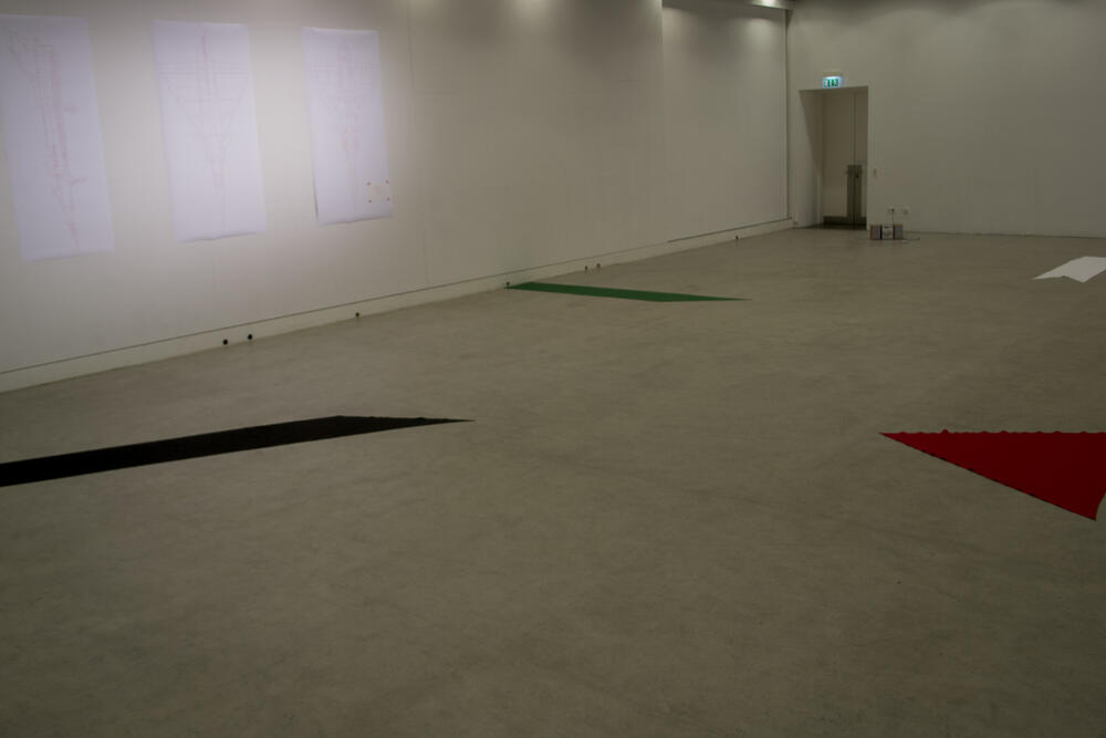 A spacious, minimalist gallery space with concrete flooring and white walls. Colored geometric shapes—black, green, and red lines or triangles—are arranged across the floor, with a projection screen visible on the left wall and an exit sign in the distance.