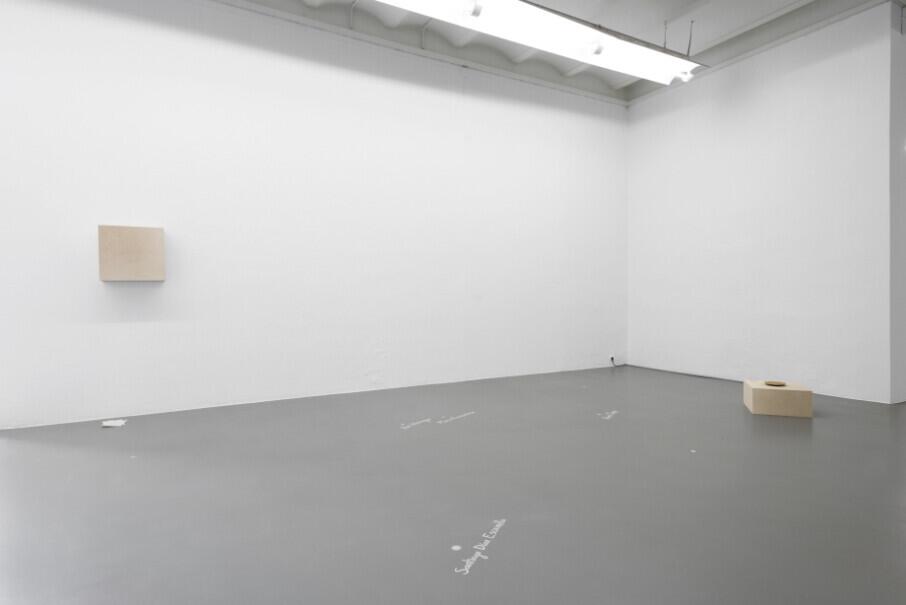 A minimalist gallery space with white walls and gray concrete floor contains three small beige wooden blocks positioned on the wall and floor, with faint markings visible on the ground surface.