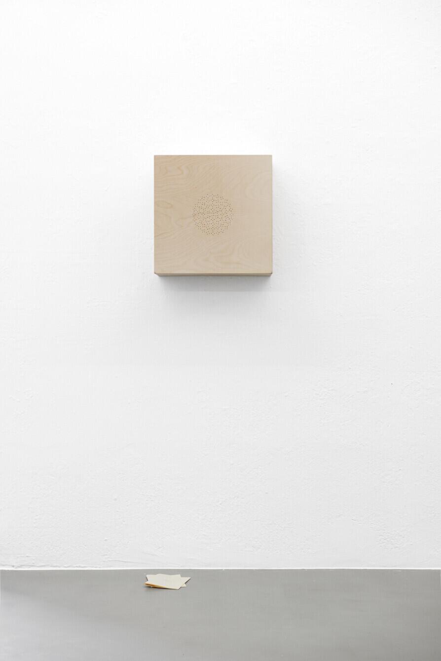 A beige square canvas with a faint circular geometric design hangs on a white wall, casting a subtle shadow. Below it, a small white object rests on a gray horizontal surface. The minimalist composition emphasizes negative space and geometric abstraction.