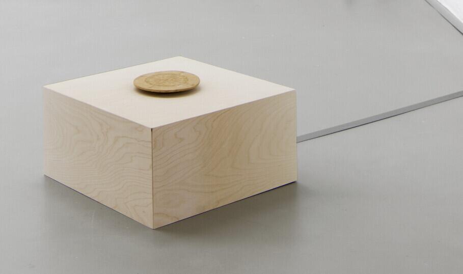 A light wooden cube sits on a gray surface with a brass circular knob mounted on its top surface, connected to a gray cable extending to the right.