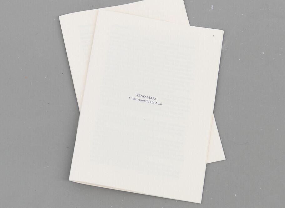 Two cream-colored folded papers or pamphlets stacked at an angle on a gray surface, with small text visible on the front cover that reads “XENO-MAPA” and "Reconstruyendo Un Atlas.