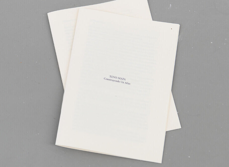 Two cream-colored folded papers or pamphlets stacked at an angle on a gray surface, with small text visible on the front cover that reads “XENO-MAPA” and "Reconstruyendo Un Atlas.